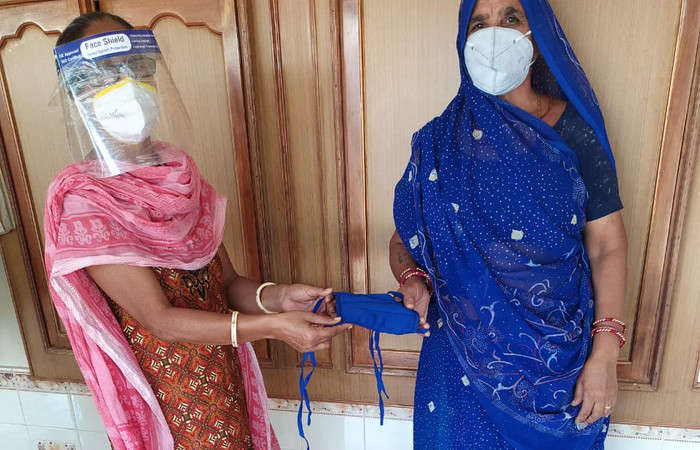 Una mujer en la India con una mascarilla y un protector facial entregando una mascarilla de tela a otra mujer que también usa una mascarilla